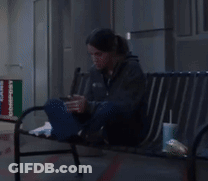 A Woman Is Sitting On A Bench Looking At Her Cell Phone GIF