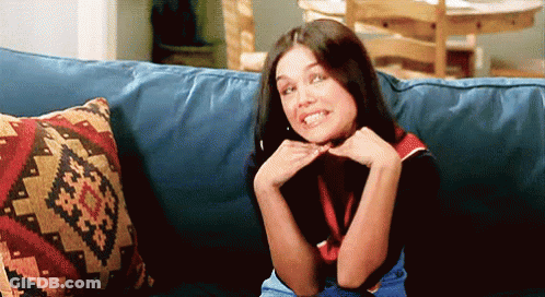 A Woman Is Sitting On A Blue Couch With A Patterned Pillow Behind Her GIF