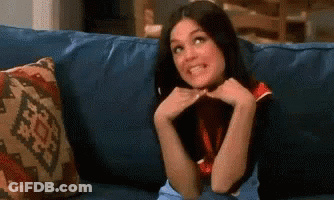 A Woman Is Sitting On A Blue Couch With Her Hands On Her Face And Smiling . GIF