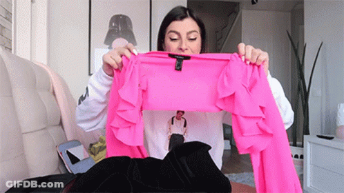 A Woman Is Sitting On A Couch Holding A Bright Pink Ruffled Top . GIF