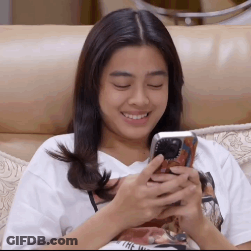 A Woman Is Sitting On A Couch Looking At Her Phone And Smiling GIF