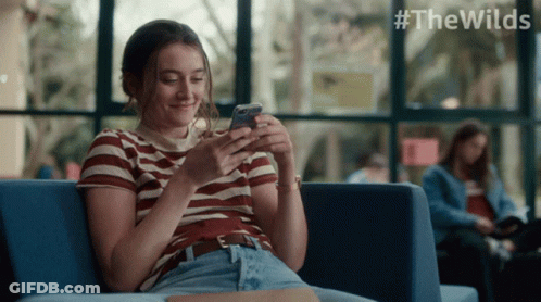 A Woman Is Sitting On A Couch Looking At Her Phone With #Thewilds Written In The Corner GIF