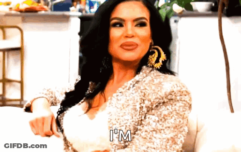 A Woman Is Sitting On A Couch Wearing A Sequined Jacket And Earrings And Saying `` I 'M '' . GIF