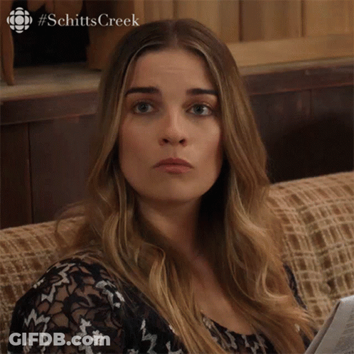 A Woman Is Sitting On A Couch With A Newspaper In Her Hand And The Word Schitts Creek On The Bottom GIF