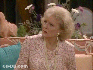 A Woman Is Sitting On A Couch With Flowers In The Background And Looking At The Camera . GIF