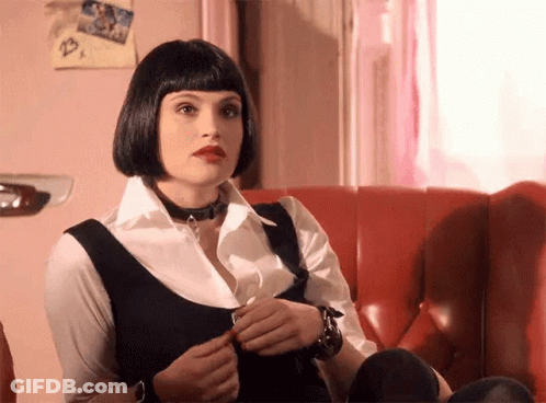 A Woman Is Sitting On A Red Couch Wearing A Choker And A Wig . GIF