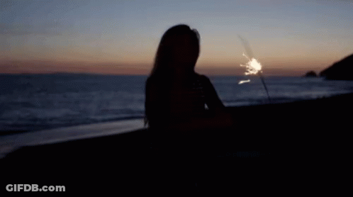 A Woman Is Sitting On The Beach Holding A Sparkler At Sunset . GIF