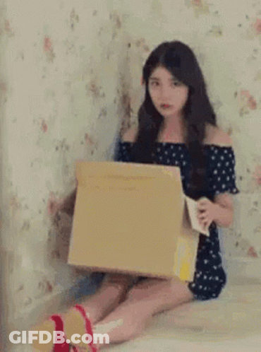 A Woman Is Sitting On The Floor Holding A Cardboard Box And A Bag . GIF