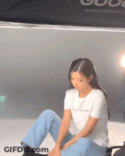 A Woman Is Sitting On The Floor Wearing A White T-Shirt That Says Calvin Klein Jeans . GIF