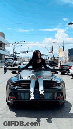 A Woman Is Sitting On The Hood Of A Sports Car GIF