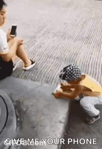 A Woman Is Sitting On The Sidewalk Next To A Child Who Is Kneeling Down And Playing A Game On His Phone . GIF