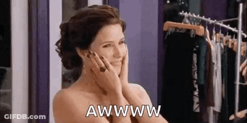 A Woman Is Smiling And Holding Her Face In Front Of A Clothes Rack In A Store . GIF
