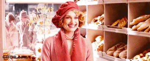 A Woman Is Smiling In A Bakery While Wearing A Red Hat And A Pink Coat . GIF