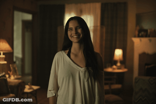 A Woman Is Smiling In A Dark Room With A Lamp In The Background GIF