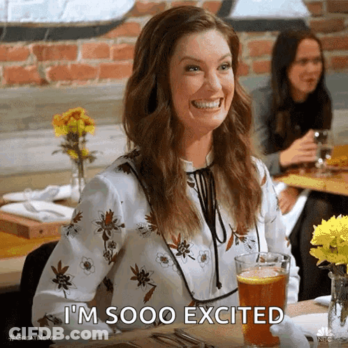 A Woman Is Smiling While Sitting At A Table With A Glass Of Beer And Says I 'M Sooo Excited . GIF