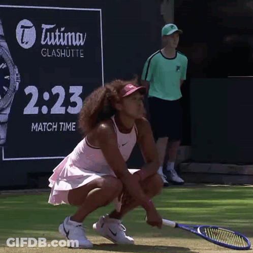 A Woman Is Squatting Down With A Tennis Racquet In Front Of An Advertisement For Tutima Glashutte GIF