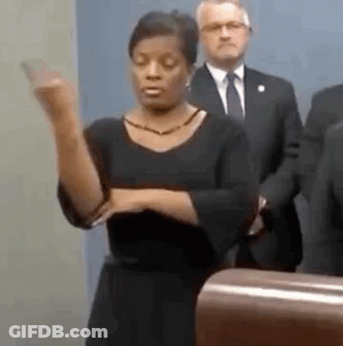 A Woman Is Standing At A Podium Giving A Speech While A Man In A Suit Stands Behind Her . GIF