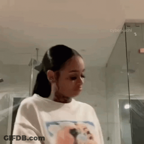 A Woman Is Standing In A Bathroom Wearing A White T-Shirt And A Ponytail . GIF