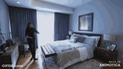 A Woman Is Standing In A Bedroom With A Large Bed And A Bench . GIF
