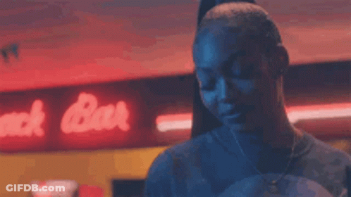 A Woman Is Standing In A Dark Room In Front Of A Neon Sign That Says Snack Bar . GIF