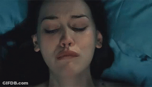 A Woman Is Standing In A Dark Room Looking Out A Window GIF