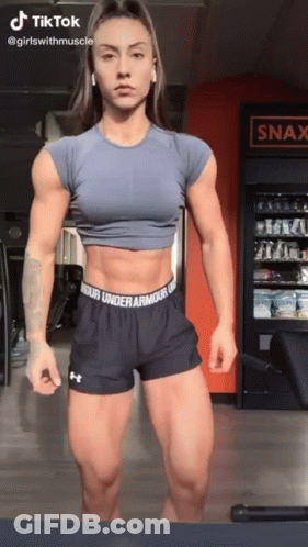 A Woman Is Standing In A Gym Wearing Under Armour Shorts And A Blue Crop Top . GIF