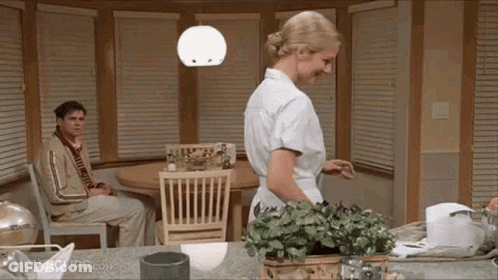 A Woman Is Standing In A Kitchen With A Man Sitting At A Table . GIF