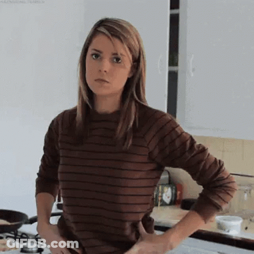 A Woman Is Standing In A Kitchen With Her Hands On Her Hips . She Is Wearing A Brown Striped Shirt . GIF