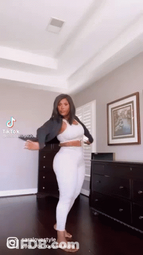 A Woman Is Standing In A Living Room Wearing White Pants And A Black Jacket . GIF
