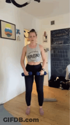 A Woman Is Standing In A Room Holding Two Dumbbells In Her Hands . GIF