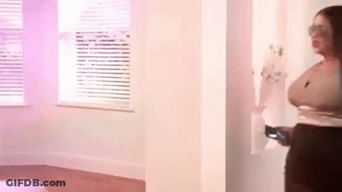 A Woman Is Standing In A Room Next To A Window Holding A Remote Control . GIF