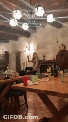 A Woman Is Standing In A Room With A Table And A Fireplace GIF