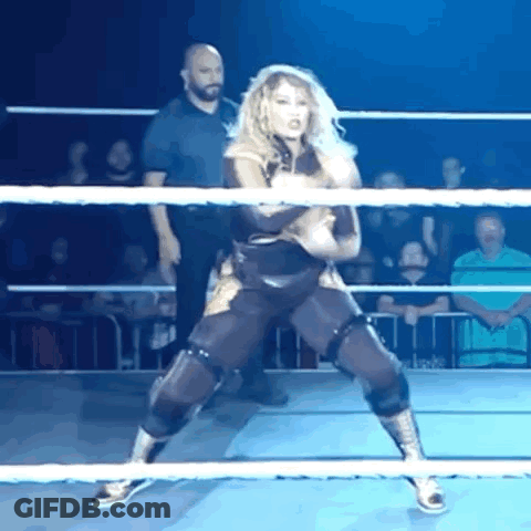A Woman Is Standing In A Wrestling Ring With A Referee In The Background GIF