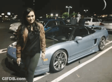 A Woman Is Standing In Front Of A Blue Sports Car With A License Plate That Says A GIF