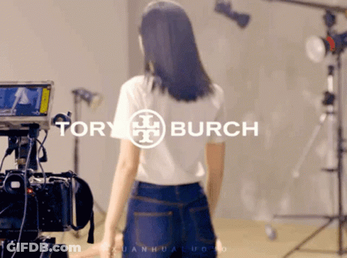 A Woman Is Standing In Front Of A Camera With Tory Burch Written On It GIF