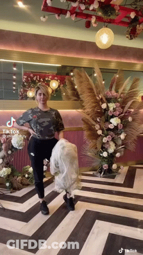 A Woman Is Standing In Front Of A Floral Arrangement In A Room With A Striped Floor . GIF
