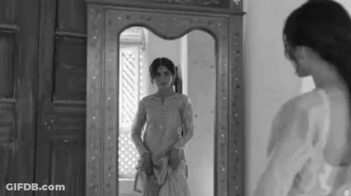 A Woman Is Standing In Front Of A Mirror Looking At Her Reflection . GIF