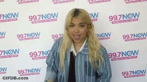 A Woman Is Standing In Front Of A Wall That Says 99.7 Now GIF