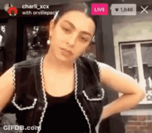 A Woman Is Standing In Front Of A Window Wearing A Black Vest And Earrings . GIF