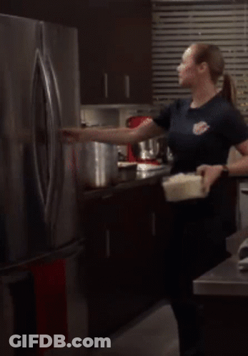 A Woman Is Standing In Front Of An Open Refrigerator Holding A Container GIF
