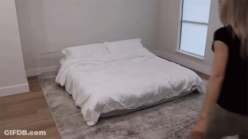 A Woman Is Standing Next To A Bed With White Sheets And Pillows GIF