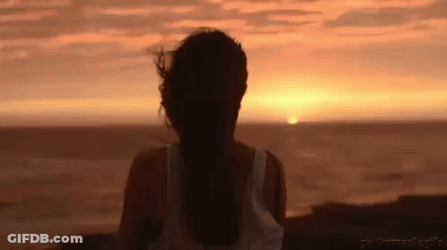 A Woman Is Standing On A Beach Watching The Sunset Over The Ocean . GIF