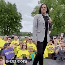 A Woman Is Standing On A Stage Holding A Microphone In Front Of A Crowd Of People Wearing Yellow Shirts . GIF