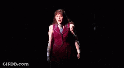 A Woman Is Standing On A Stage In A Dark Room Wearing A Red Vest And Tie . GIF