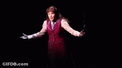 A Woman Is Standing On A Stage In A Dark Room Wearing A Red Vest And Tie . GIF