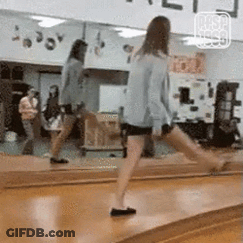 A Woman Is Standing On A Wooden Floor In Front Of A Large Mirror . GIF