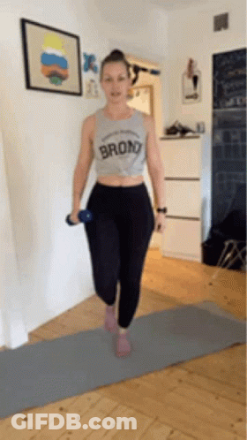A Woman Is Standing On A Yoga Mat Holding A Dumbbell And Wearing A Shirt That Says Bronx . GIF