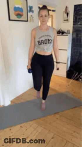 A Woman Is Standing On A Yoga Mat Holding A Dumbbell And Wearing A Shirt That Says Brooklyn . GIF