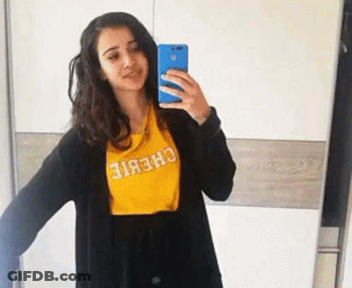 A Woman Is Taking A Selfie In Front Of A Mirror Wearing A Yellow Chebie Shirt GIF