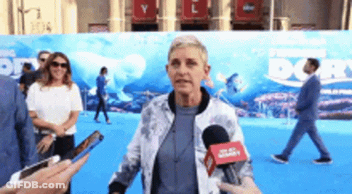 A Woman Is Talking Into A Microphone While Standing In Front Of A Sign That Says Finding Dory . GIF